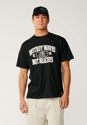 Hombre con gorra negra y camiseta negra con el texto y gráfico ""Destroy Waves Not Beaches"", de pie contra un fondo claro y liso.