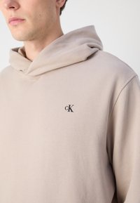 Beige kapucnis pulóver puha anyagból, nagy elülső zsebbel. A mellkasán fekete "cK" logóval és tágas kapucnival rendelkezik.
