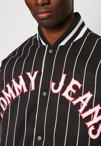 Mustad must jacket valgete vertikaalsete triipudega. Omab punaseid broderitud "TOMMY JEANS" tekste, nupuvajutusi ja ribaga kraed.