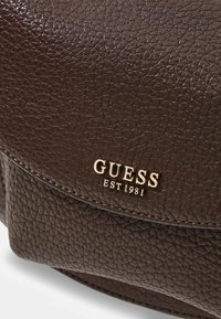 Bruine leren tas met een textuur en een goudkleurig "GUESS EST 1981" logo aan de voorkant, met een gebogen klep en zichtbare stikdetails.
