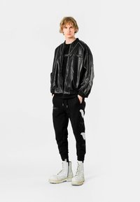 Chaqueta bomber de cuero negra con cierre de cremallera, acentos a rayas, combinada con pantalones deportivos negros con adornos blancos y botas blancas gruesas.