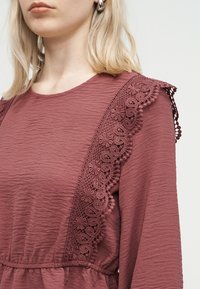 Γυναίκα που φορά ένα φορεμα σε μωβ απόχρωση με υφή και δαντέλα με λουλούδια στους ώμους, καθώς και ασημένια σκουλαρίκια κρίκους, εμφανίζεται από το πηγούνι μέχρι τη μέση.