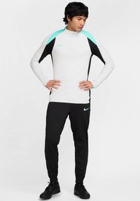 Herren Nike Sportbekleidung mit einem weißen Langarmoberteil mit türkisfarbenen und schwarzen Akzenten, kombiniert mit schwarzen Hosen. Texturiertes Material, schlanker Schnitt.