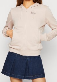 Rosa zip-up hoodie med framficka, justerbar huva och subtil logotyp. Matcher med en mörk denimfaldskjol.