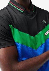 Man som bär en Lacoste sporttröja med svart topp, grön mitten, blå botten och logpatchar på bröstet och kragen.