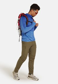 Maglia blu a maniche lunghe con zip, pantaloni beige con zip e scarpe sportive grigie. Zaino rosso e nero con vari cinturini, indossato a spalla.