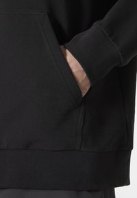 Sudadera negra con bolsillo tipo canguro, puños y dobladillo acanalados. Hecha de tela suave, con costuras visibles a lo largo de las costuras.