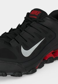 Μαύρο αθλητικό παπούτσι Nike με ασημί λογότυπο swoosh, κόκκινη ετικέτα "NIKE TRAINING" στη γλώσσα και κόκκινα στοιχεία απορρόφησης κραδασμών στη σόλα.