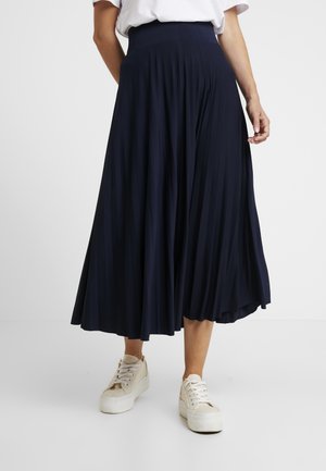 Vrouw draagt een marineblauwe geplooide midirok en witte platform sneakers, staand tegen een effen lichte achtergrond.