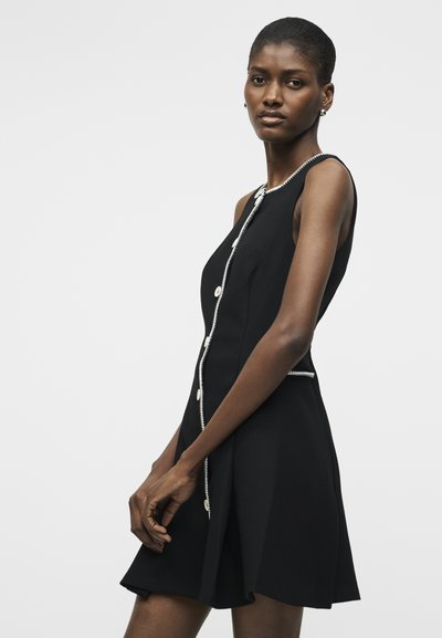 Γυναίκα με κοντά μαλλιά που φοράει μαύρο φόρεμα χωρίς μανίκια με άσπρες ρίγες και κουμπιά, στέκεται μπροστά σε απλό ανοιχτόχρωμο φόντο.