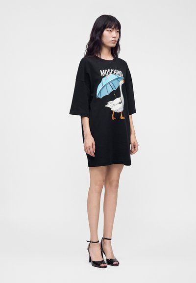 Γυναίκα που φοράει μαύρο oversized φόρεμα T-shirt με γραφικό μιας χήνας που κρατάει μια μπλε ομπρέλα και τη λέξη Moschino.