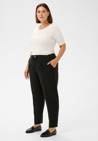 O femeie stând cu o mână în buzunar, îmbrăcată cu un top alb cu mânecă scurtă, pantaloni negri tapered și mocasini negri, pe un fundal simplu.