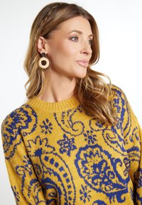 Geel gebreid sweater met blauwe paisleypatronen, ronde halslijn en textiel stof. Grote ronde oorbellen maken de look compleet.