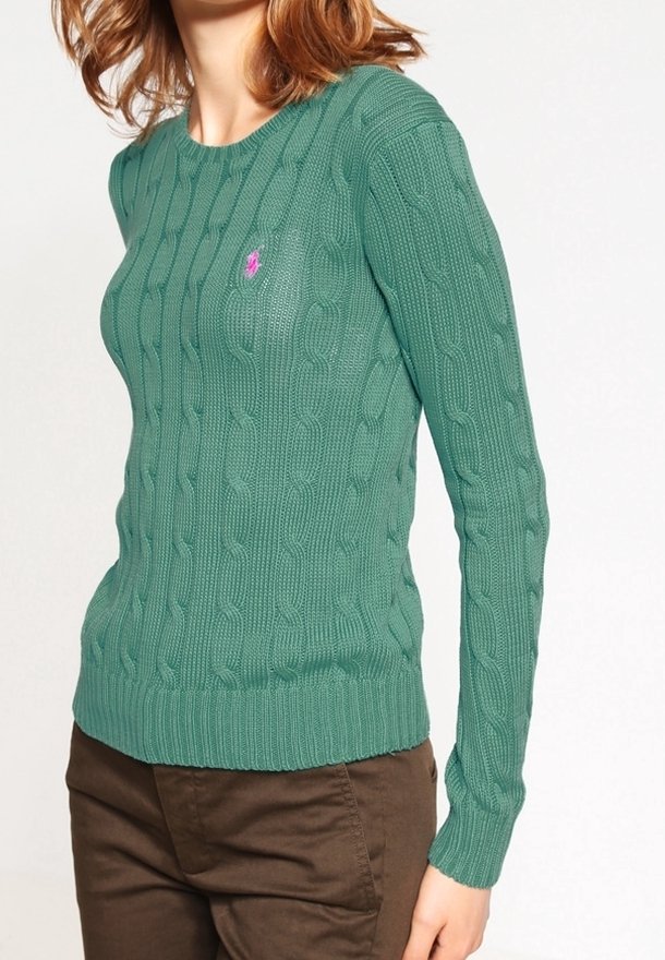 Femme portant un pull vert en maille torsadée avec un petit logo rose brodé sur la poitrine et un pantalon marron, sur un fond uni.