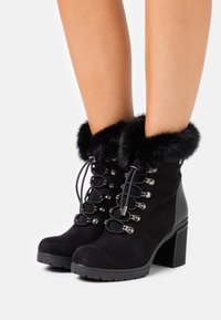 Stivali alla caviglia in suede nero con rifiniture in pelliccia sintetica, lacci anteriori, dettagli in metallo argentato e tacco spesso. Suola texturizzata per una maggiore aderenza.