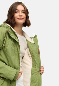 Grön vadderad jacka med en plysch beige foder, som har en huva, dragsko och metallaccenter. Öppen, som visar inredningens textur och design.