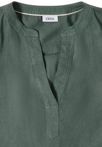 Haut en lin vert avec une encolure fendue et une bordure cousue, avec une étiquette blanche portant le nom de la marque « CECIL » à l’intérieur du col.
