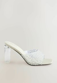 Mules blanches avec un dessus orné de cristaux et un talon bloc transparent. Semelle plate avec texture lisse et design à bout ouvert.