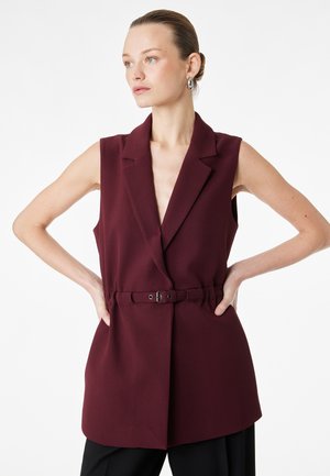 Donna con i capelli raccolti in uno chignon, che indossa un blazer senza maniche color bordeaux scuro con cintura e orecchini a cerchio d'argento, in piedi con le mani sui fianchi.