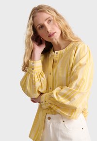 Camicia a righe gialle con maniche a sbuffo e scollatura tonda, caratterizzata da righe chiare e scure, abbinata a pantaloni bianchi.