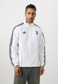 Hvid atletisk jakke med sorte striber på ærmerne, grønne accenter og lynlås lukning. Har Juventus logo og røde side detaljer.