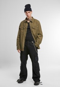 Jachetă verde măslinie cu nasturi, cu două buzunare pe piept, peste un top negru, asortată cu pantaloni cargo negri, dotati cu multiple buzunare și un lanț.