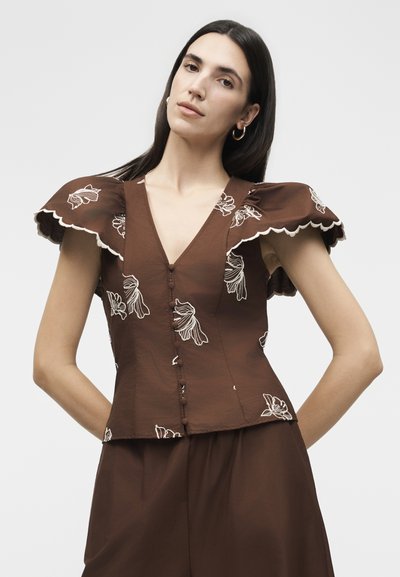 Γυναίκα που φορά καφέ μπλούζα με κουμπιά, μεγάλες φλοράλες πιέτες στα μανίκια και λευκή κεντητή διακόσμηση λουλουδιών, συνδυασμένη με καφέ παντελόνι που ταιριάζει.