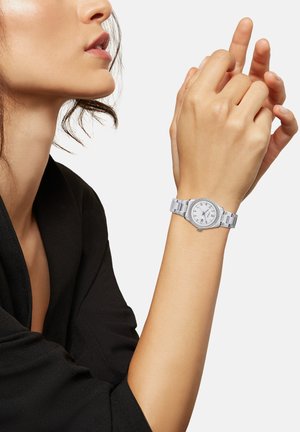 Montre en métal argenté avec un cadran blanc rond, des marqueurs d'heure allongés et un bracelet à maillons. Portée sur une main contre un fond en tissu noir.