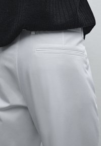 Hvide bukser med en glat overflade, der har en vandret baglomme med en syning rundt om, kombineret med en mørk struktureret sweater.