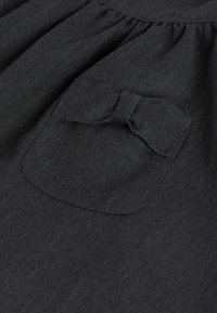 Robe en tissu noir avec un corsage froncé et une poche carrée ornée d'un nœud, mettant en valeur une texture lisse et un design structuré.