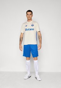 Polo beige avec des accents bleus et un logo, associé à un short bleu et des chaussettes blanches. Des chaussures de football blanches et noires.