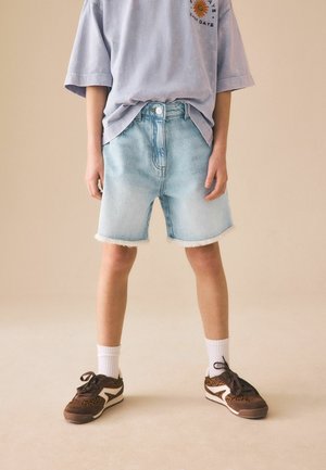 Dziecko noszące jasnoniebieskie dżinsowe szorty, luźny lawendowy T-shirt z grafiką słonecznika, białe skarpetki i brązowe trampki w panterkę.