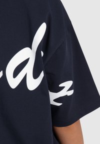 Tmavě modré bavlněné tričko s velkým bílým potiskem na přední straně a menším bílým designem na rukávu. Krátké rukávy a volný střih.