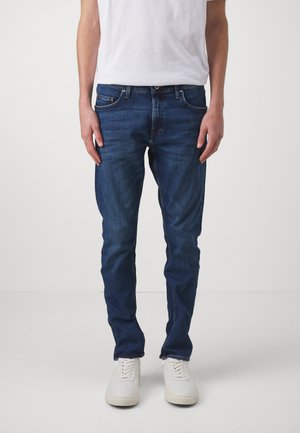 Blue denim jeans with a slim fit, featuring a medium wash, subtle fading, five pockets, and a button closure. Paired with white sneakers.