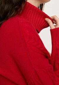 Pull rouge en maille tricotée avec un col roulé épais côtelé, présentant des motifs en maille torsadée texturée le long des manches et du corps.