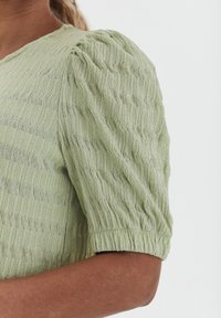Top verde chiaro con texture e maniche corte voluminose, caratterizzato da righe verticali e un polsino arricciato per una silhouette strutturata.