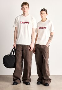 Dwie osoby stoją obok siebie, ubrane w kremowe koszulki z czerwonym logo, luźne brązowe spodnie oraz ciemne buty o zaokrąglonych noskach. Jedna trzyma czarną torbę.