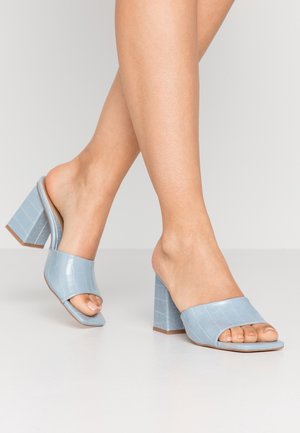 Pies con mules de tacón ancho, puntera abierta y relieve de cocodrilo azul sobre una superficie blanca, con un pie avanzando.