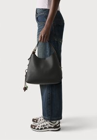 Sac hobo en cuir noir avec une silhouette incurvée, doté d'une bandoulière marron et d'un pompon décoratif avec des accents verts et bruns. Tenue décontractée avec un jean.