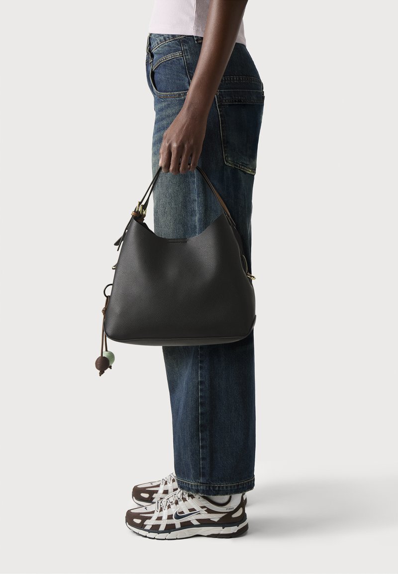 Sac hobo en cuir noir avec une silhouette incurvée, doté d'une bandoulière marron et d'un pompon décoratif avec des accents verts et bruns. Tenue décontractée avec un jean.