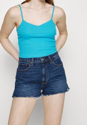 Vrouw draagt een turquoise geribd tanktop en donkerblauwe spijkershorts met hoge taille en rafelige zoom, staand met handen achter de rug.