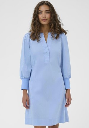 Žena s kučeravými vlasmi, oblečená v svetlomodrých dlhých rukávoch šatách s výstrihom do V a zapínaním na gombíky na manžetách, stojaca pred jednoduchým pozadím.