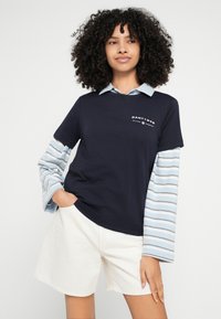 Tmavomodré tričko s svetlomodrým golierom a pruhovanými dlhými rukávmi. Bavlnená zmes, na hrudi má logo, nosené s béžovými krátkymi nohavicami.