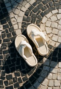 Ζευγάρι μπεζ σανδάλια με δεμένους λουρίδες σε μαύρο και άσπρο κυκλικό ψηφιδωτό δάπεδο με ηλιακό φως και σκιές.
