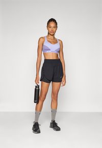 Lilla sport-BH med Nike-merke, svarte treningsshorts, grå sokker og svarte joggesko. Holder en svart vannflaske. Studiobakgrunn.
