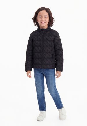 Smiling child with shoulder-length hair wearing a black patterned puffer jacket, blue jeans, and white sneakers, standing against white background.