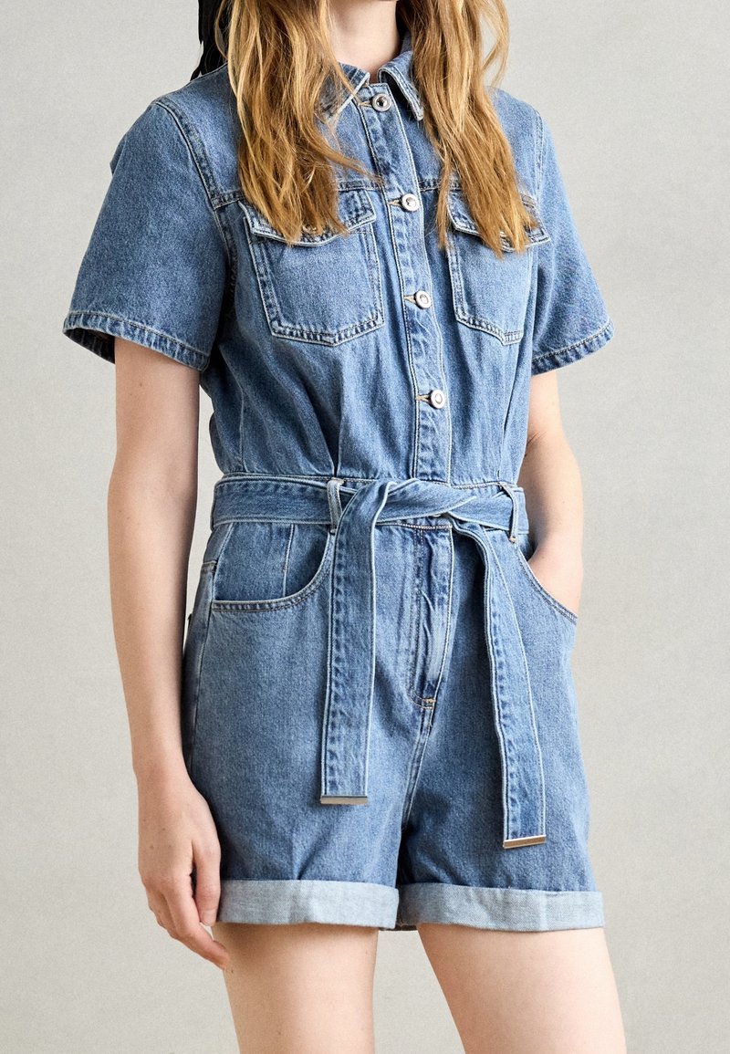 Femme portant une combinaison en denim bleu à manches courtes avec des boutons sur le devant, deux poches poitrine, un short roulotté et une ceinture nouée à la taille.