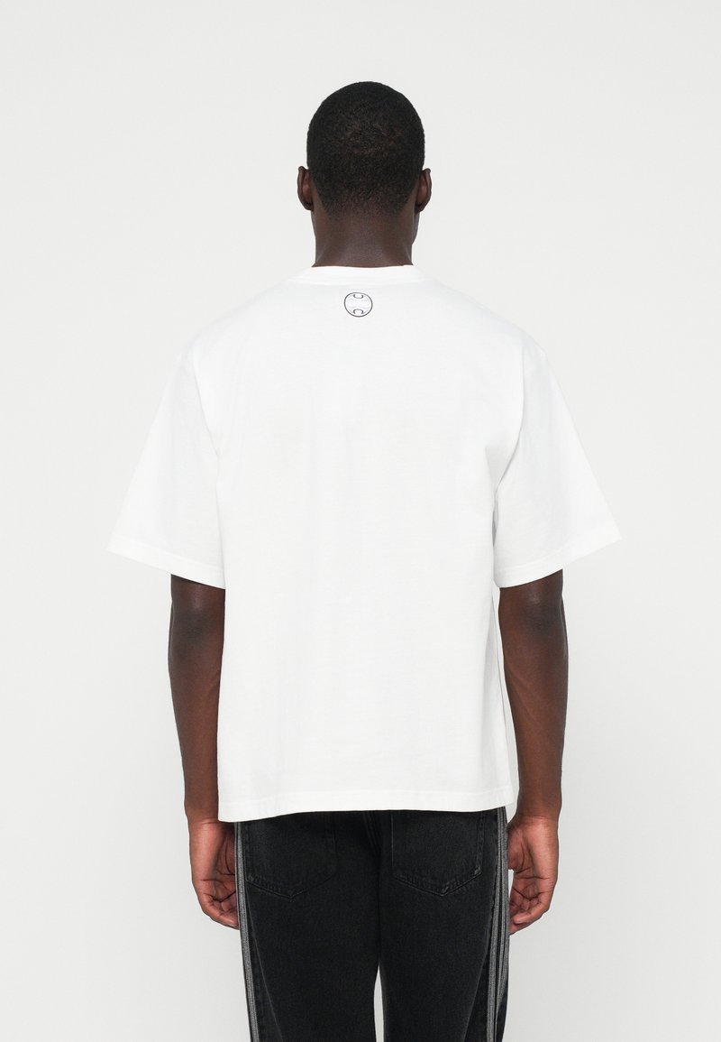 Hombre visto desde atrás, vistiendo una camiseta blanca de manga corta sin diseño, con un pequeño logotipo redondo cerca del cuello, combinada con jeans negros.