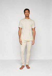 T-shirt de algodão bege e calças combinando com punhos canelados. A t-shirt apresenta um pequeno logótipo ao peito. O modelo está descalço contra um fundo simples.