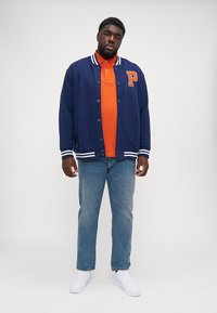 Navy varsity jacket with white stripes, orange polo shirt beneath, light blue jeans, and white sneakers. Notable "P" logo on jacket.